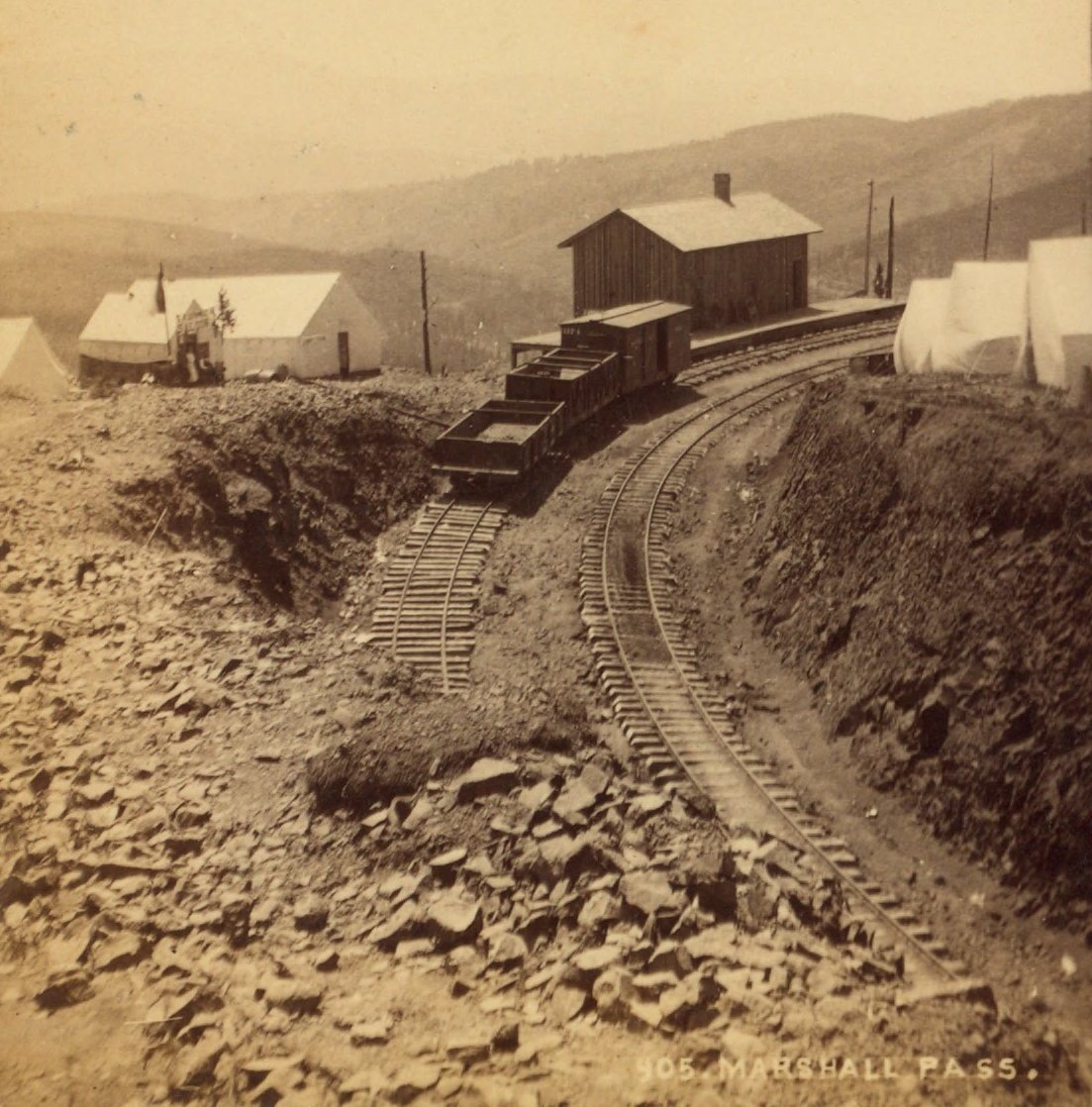 Fotografia antiga de uma ferrovia de bitola estreita, com vagões, uma estação de madeira e tendas, no alto das Montanhas Rochosas do Colorado