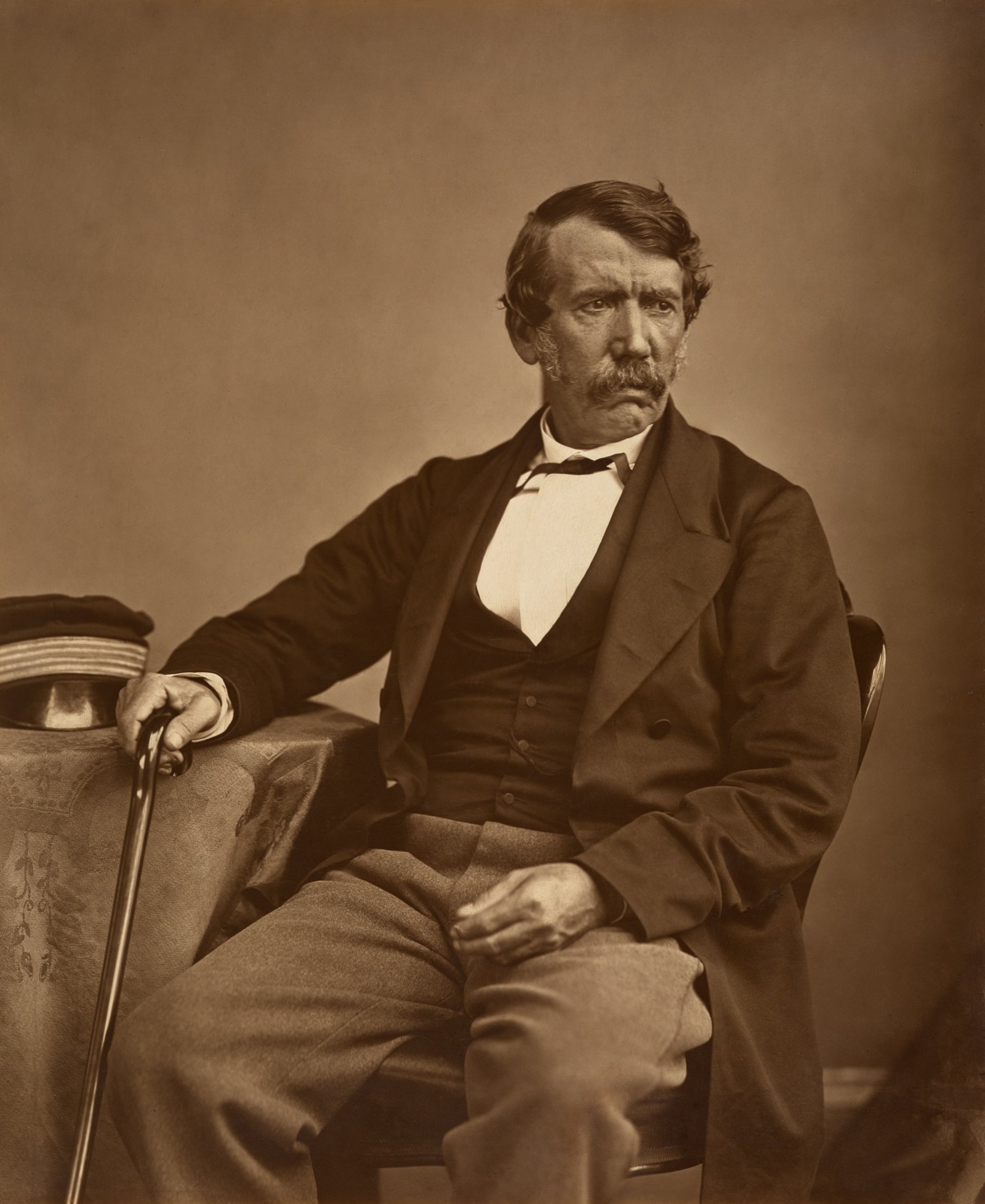 Retrato fotográfico de David Livingstone, de bigode, sentado com a bengala e o boné de viagem sobre a mesa