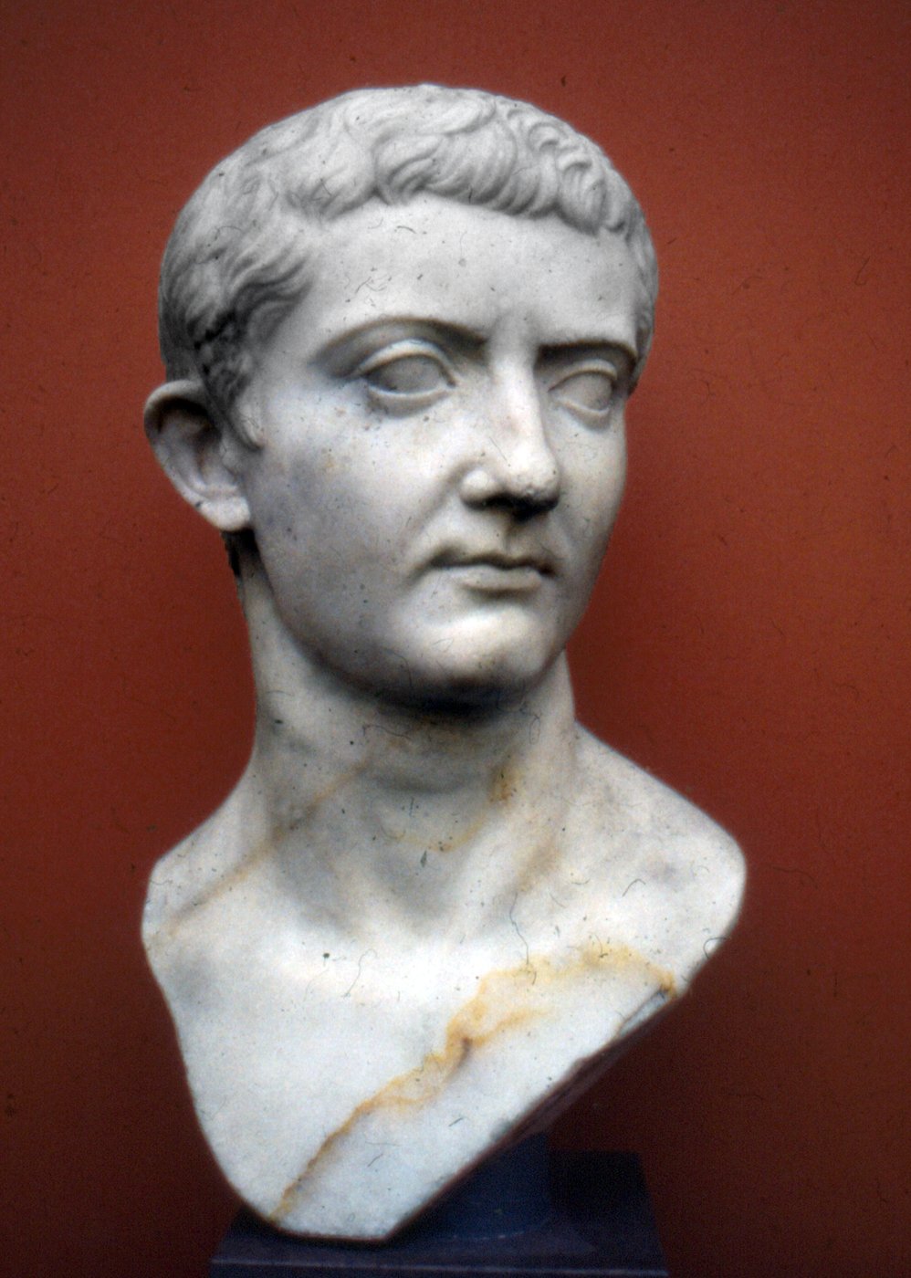 Busto de mármore do imperador romano Tibério, de rosto sério e cabelo curto em mechas sobre a testa